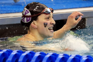 Hannah Orbach-Mandel pictured at the edge of a pool at the end of a race, smiling, wearing a swim cap and with a Kenyon shield temporary tattoo on her cheek.